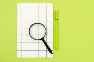 Magnifying glass on a grid-patterned notebook beside a pen on a lime-green background.